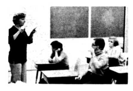 A black and white photo of a teacher talking to students seated in desks in a classroom.
